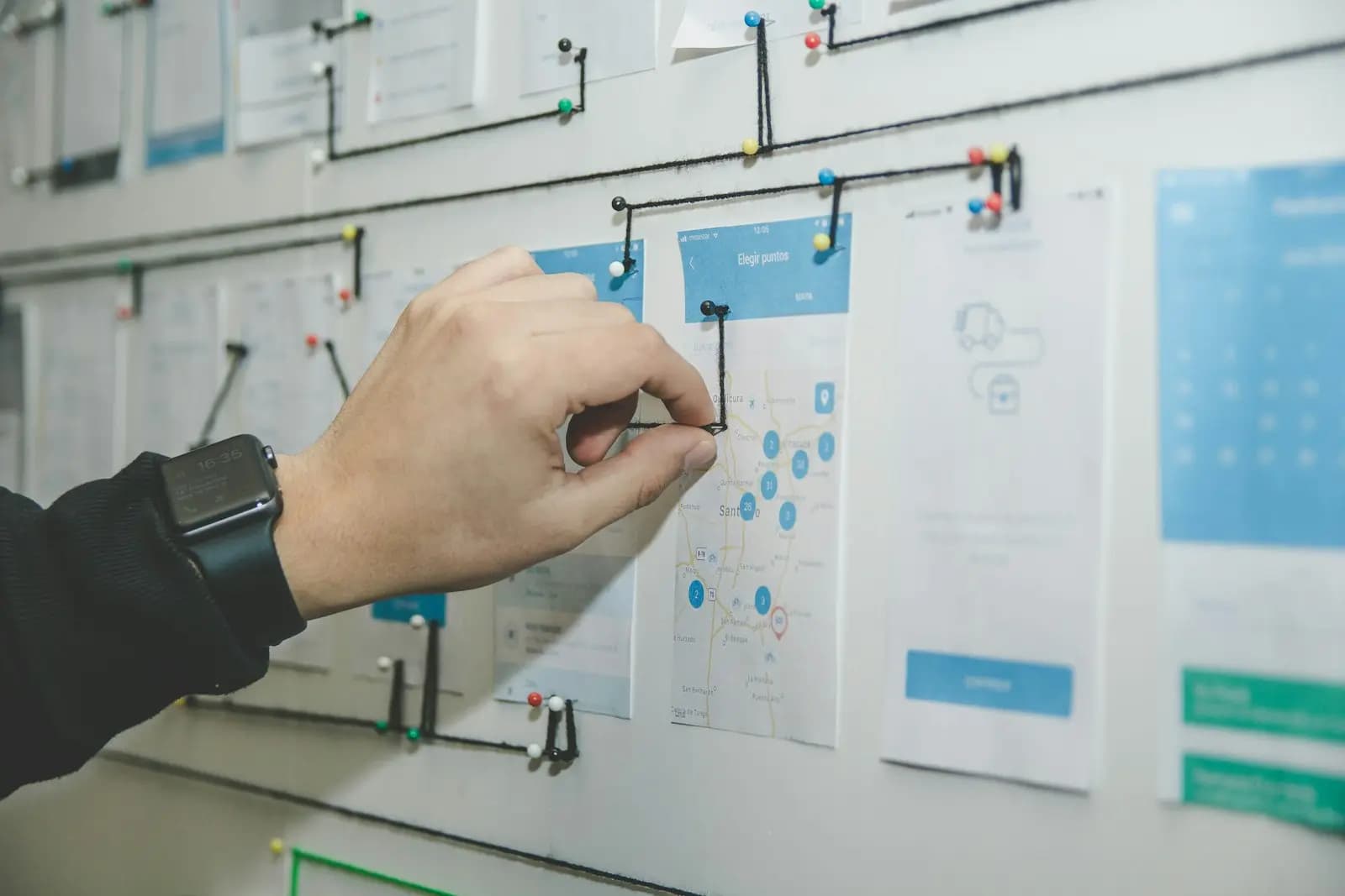 Eine Hand befestigt eine Karte an einer Wand, auf der Arbeitsschritte mit Fäden verbunden sind.
